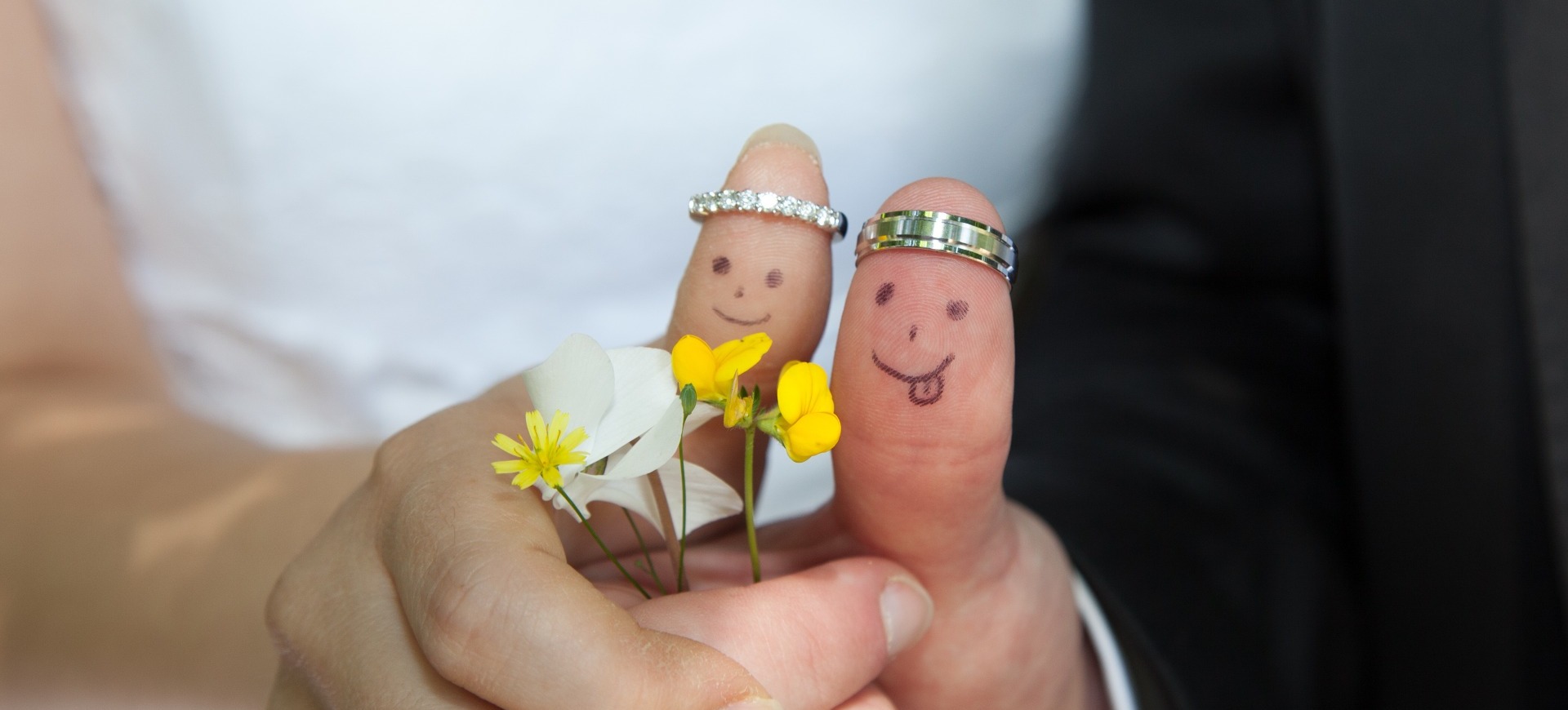 A Bride & Groom's thumbs with drawn smiley faces, holding four small flowers, wearing their wedding rings as crowns.