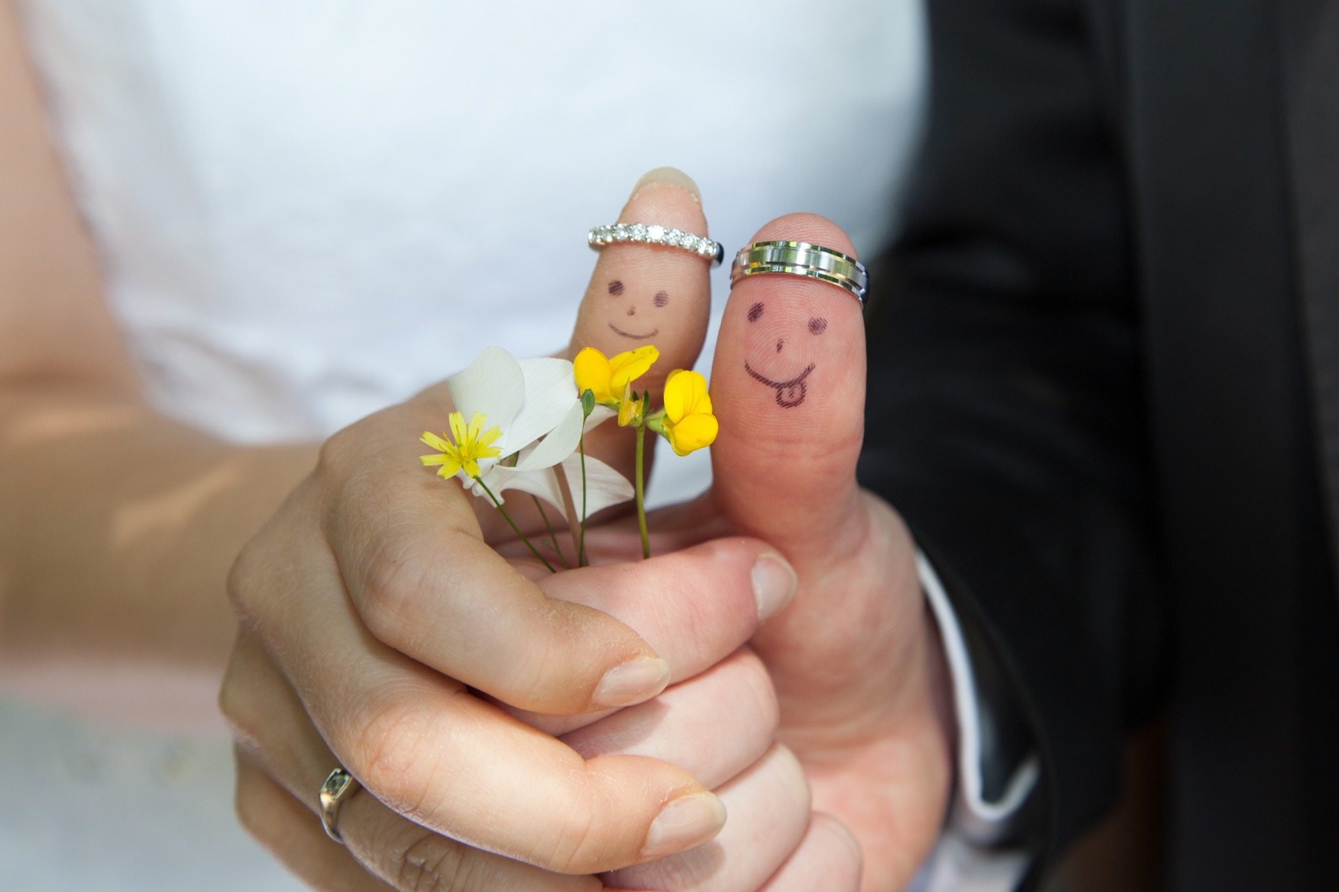 A couple holding their thumbs close together, with smiling faces drawn on, holding a few small flowers, wearing wedding rings as crowns. 