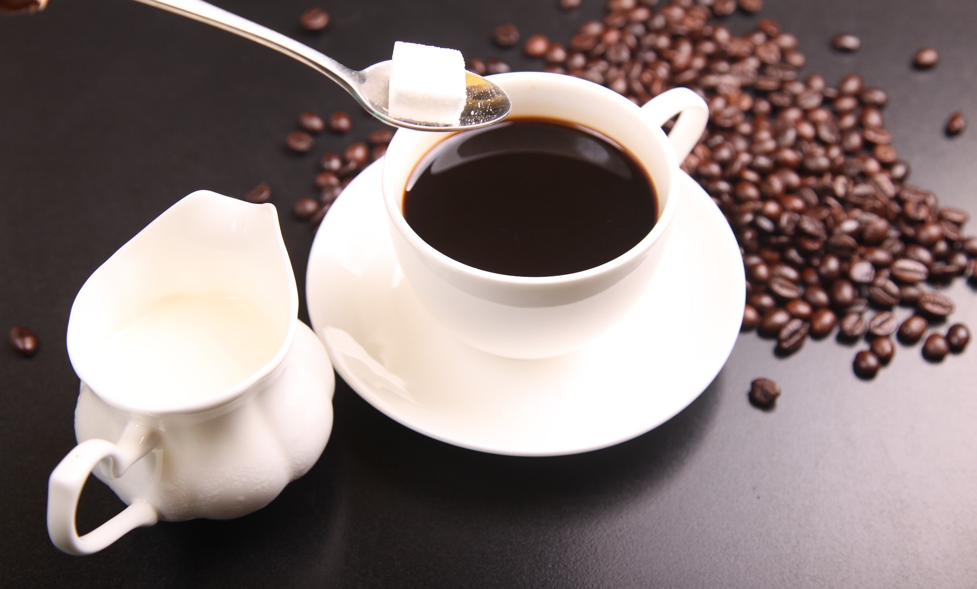 A black coffee in a cup and saucer, with coffee beans around.  A person is holding a spoon with a cube of sugar over the coffee, and there is a jug of milk to the side.  Our milk allergy solicitors are here to assist if you have been sold containing undeclared milk.