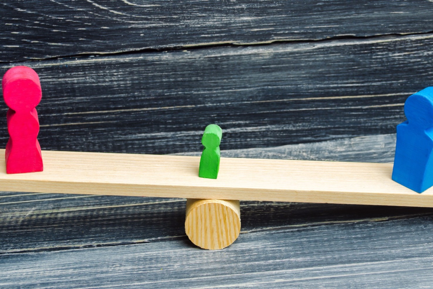 A wooden see-saw, with a pink figure on one end,a blue figure on the other, and a small green figure in the middle.