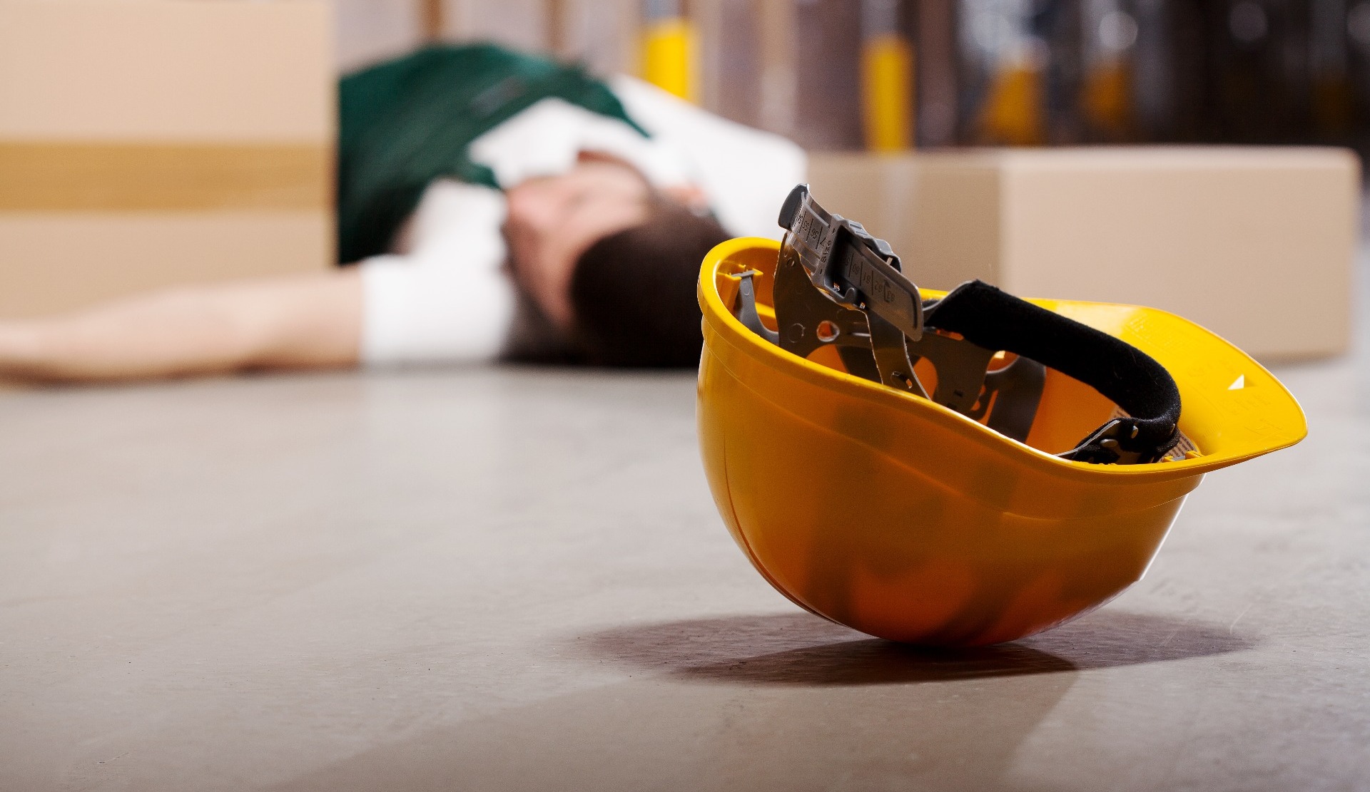A man who has been injured at work, laying on the floor.  His hard hat has rolled away from him.
