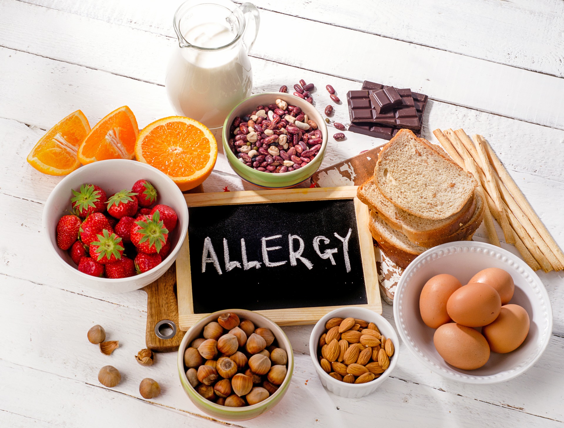 'Allergy' written on a chalk board, surrounded by allergens, including strawberries, bread, eggs, fruit, nuts and milk.