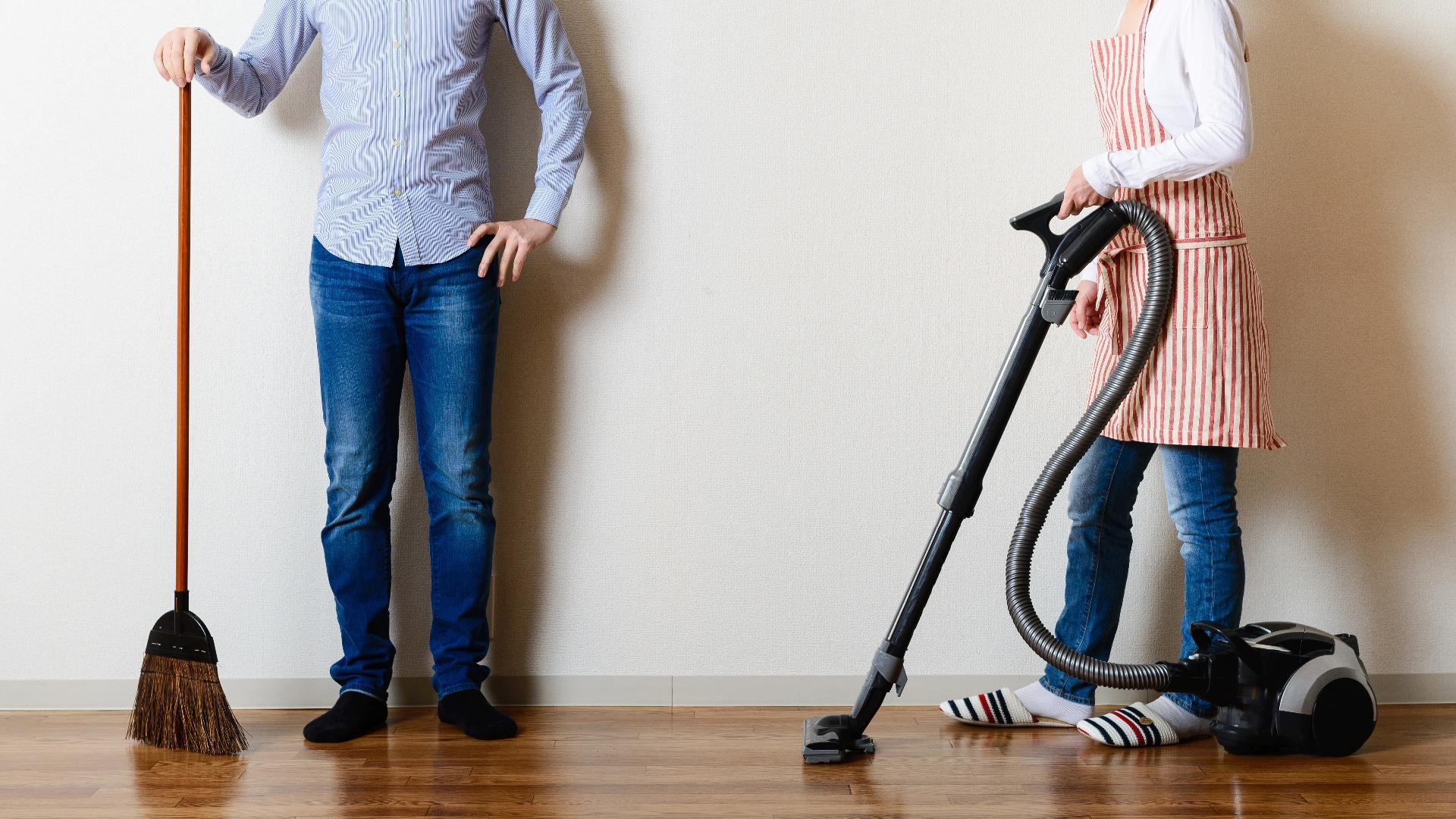 A couple, one holding a broom, and the other stood away, holding a hoover.