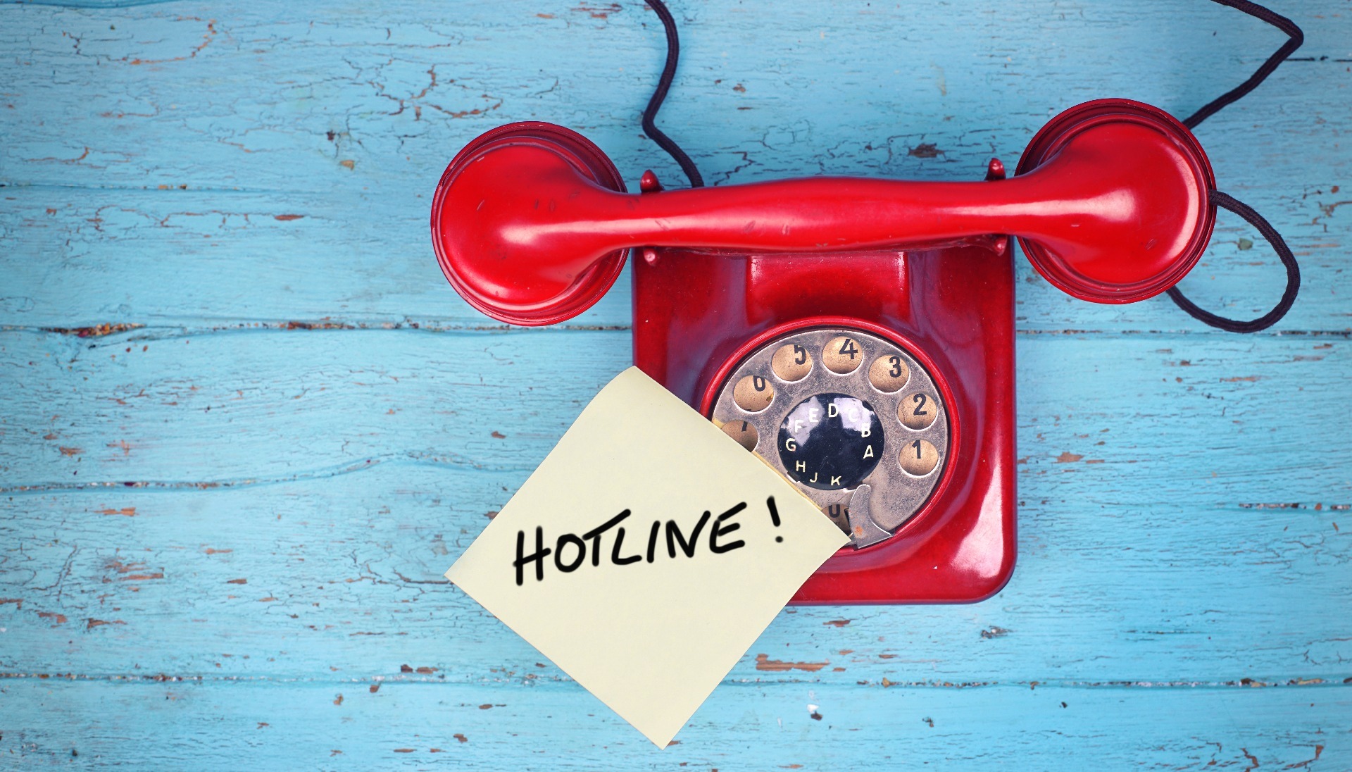 A red, dial-up old-fashioned telephone, with 'Hotline' written on a yellow sticky note, stuck to the 'phone.