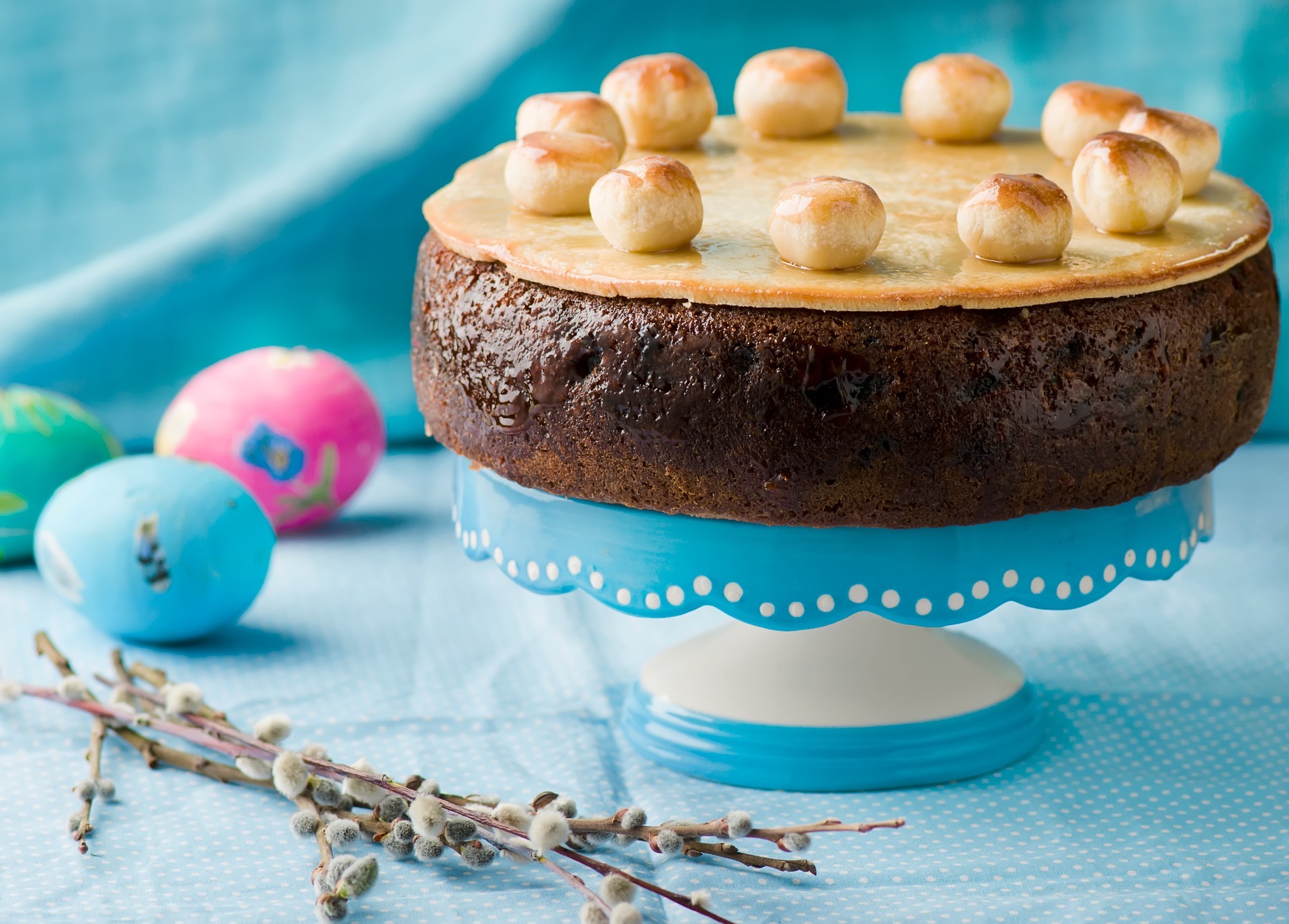 A traditional Simnel cake, on a blue and white cake stand, with a green, blue and pink faux egg and some flowers around the bottom of the stand.