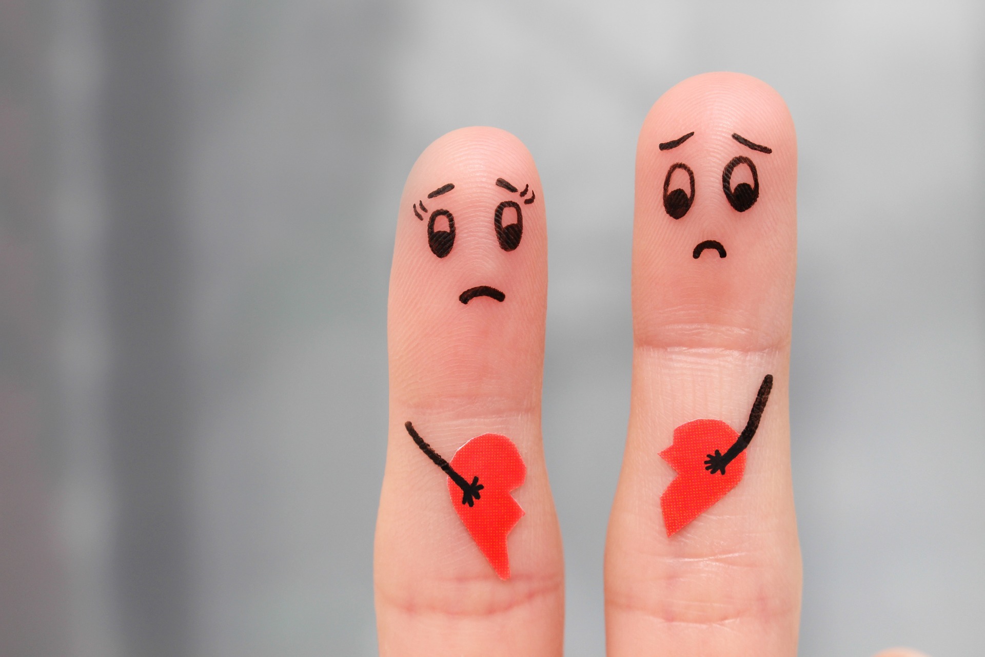 Two fingers, with a sad male face and a sad female face drawn on, each holding half of a red, broken heart.