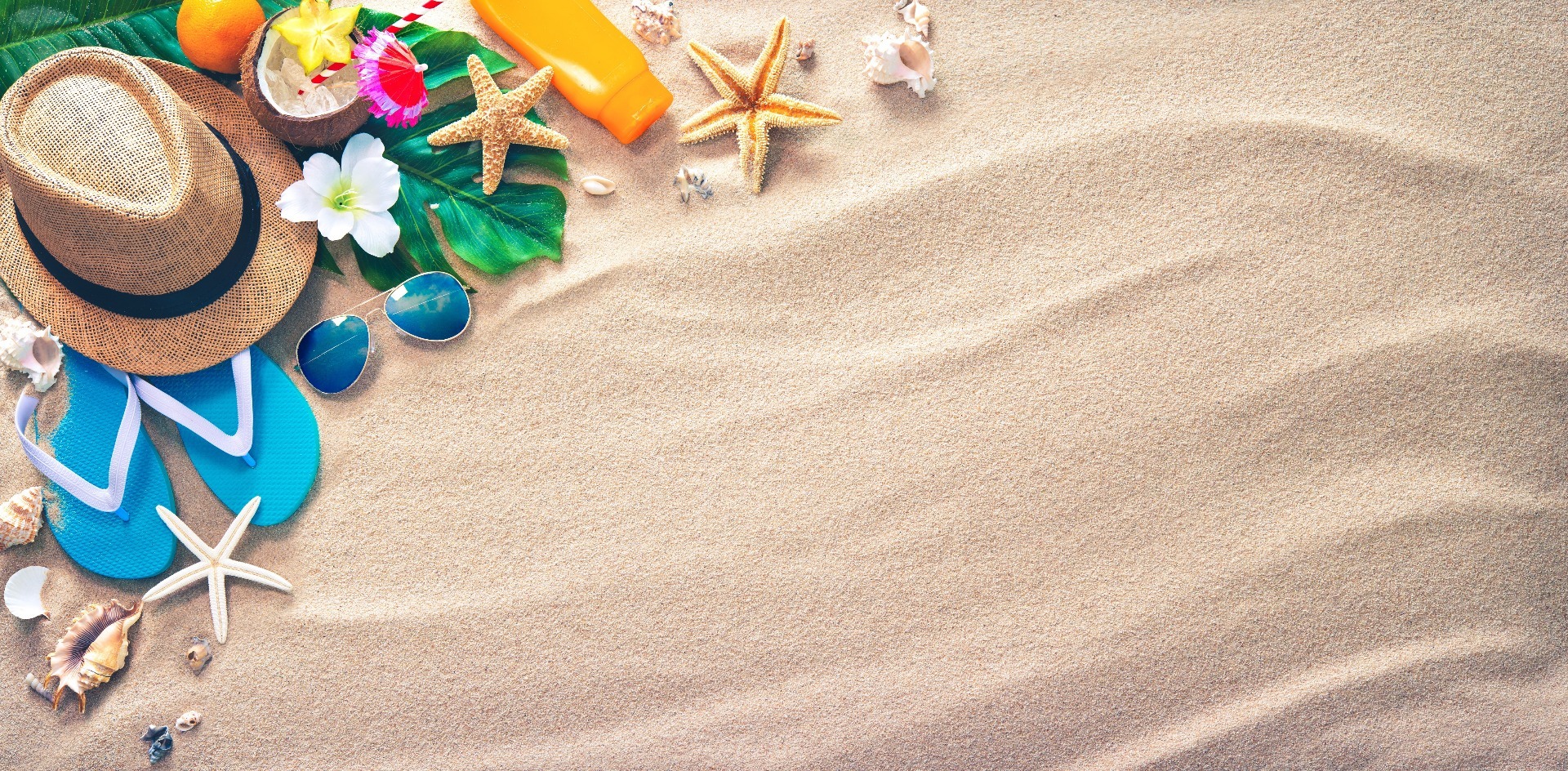 A beach of sand, with beach items placed in the top left hand corner, including a sun hat, a pair of blue flip flops, a bottle of suncream, a pair of sunglasses, a coconut and some leaves, flowers and shells.
