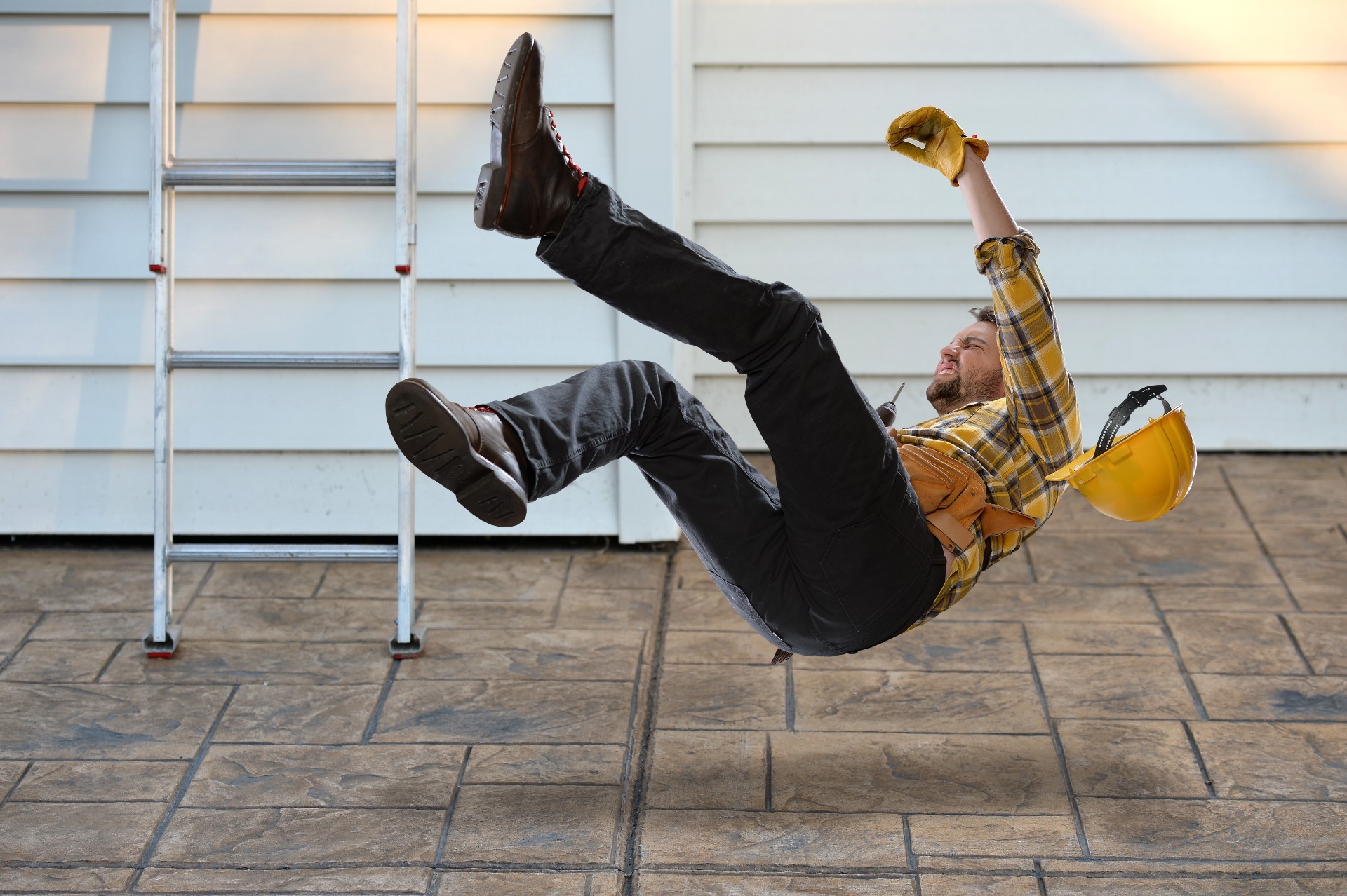 A person wearing jeans, boots and a yellow shirt, falling from a ladder, with their hard-hat falling to the floor; our No Win No Fee Solicitors can assist with making an injury compensation claim if you've been injured at work.
