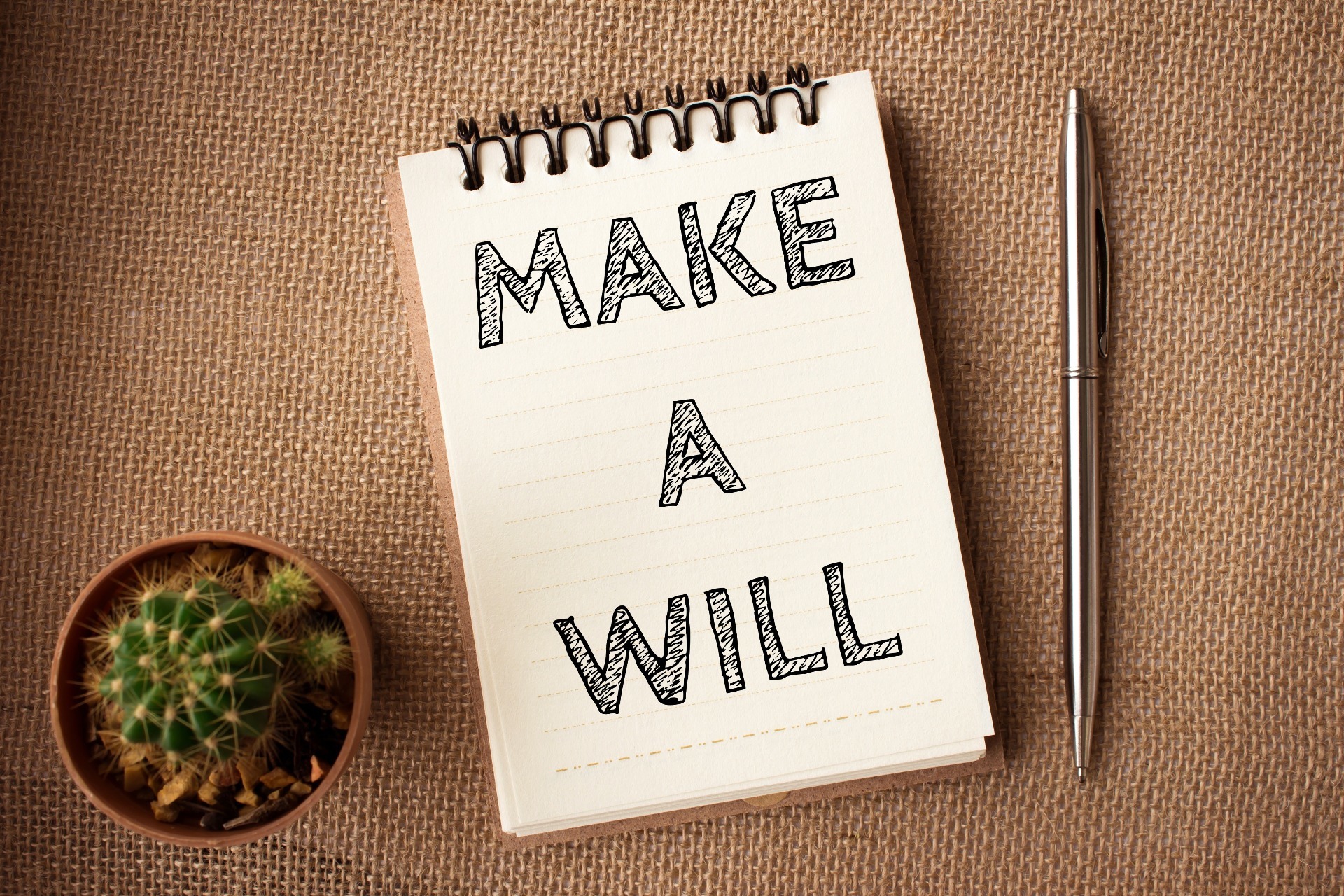 "Make a Will" written on a small notebook, against a table, with a cactus and a pen; our Wills Solicitors can assist you with the preparation of your Will all for an affordable fixed-fee.