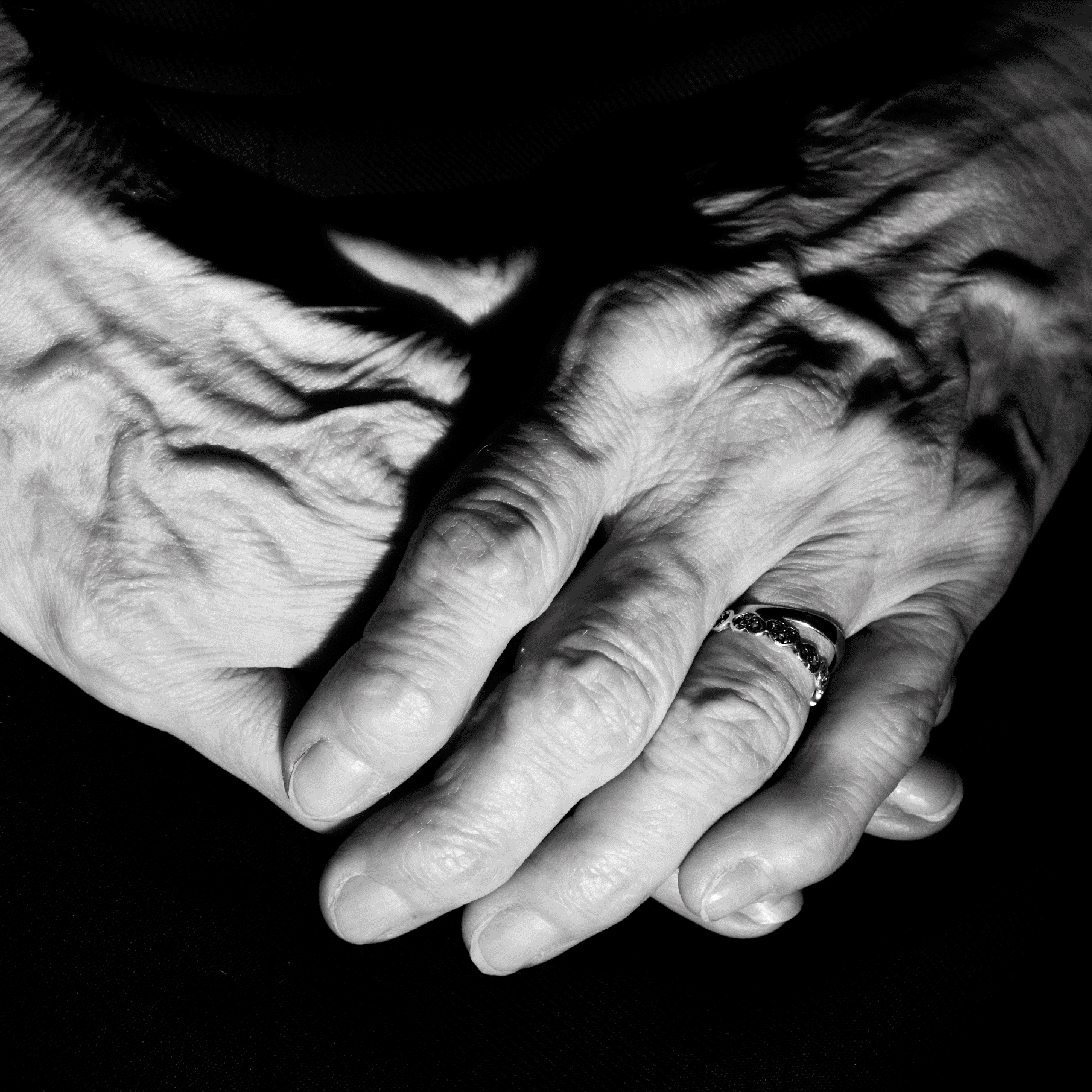 An elderley person's hands clasped together, with two rings on the left second finger.