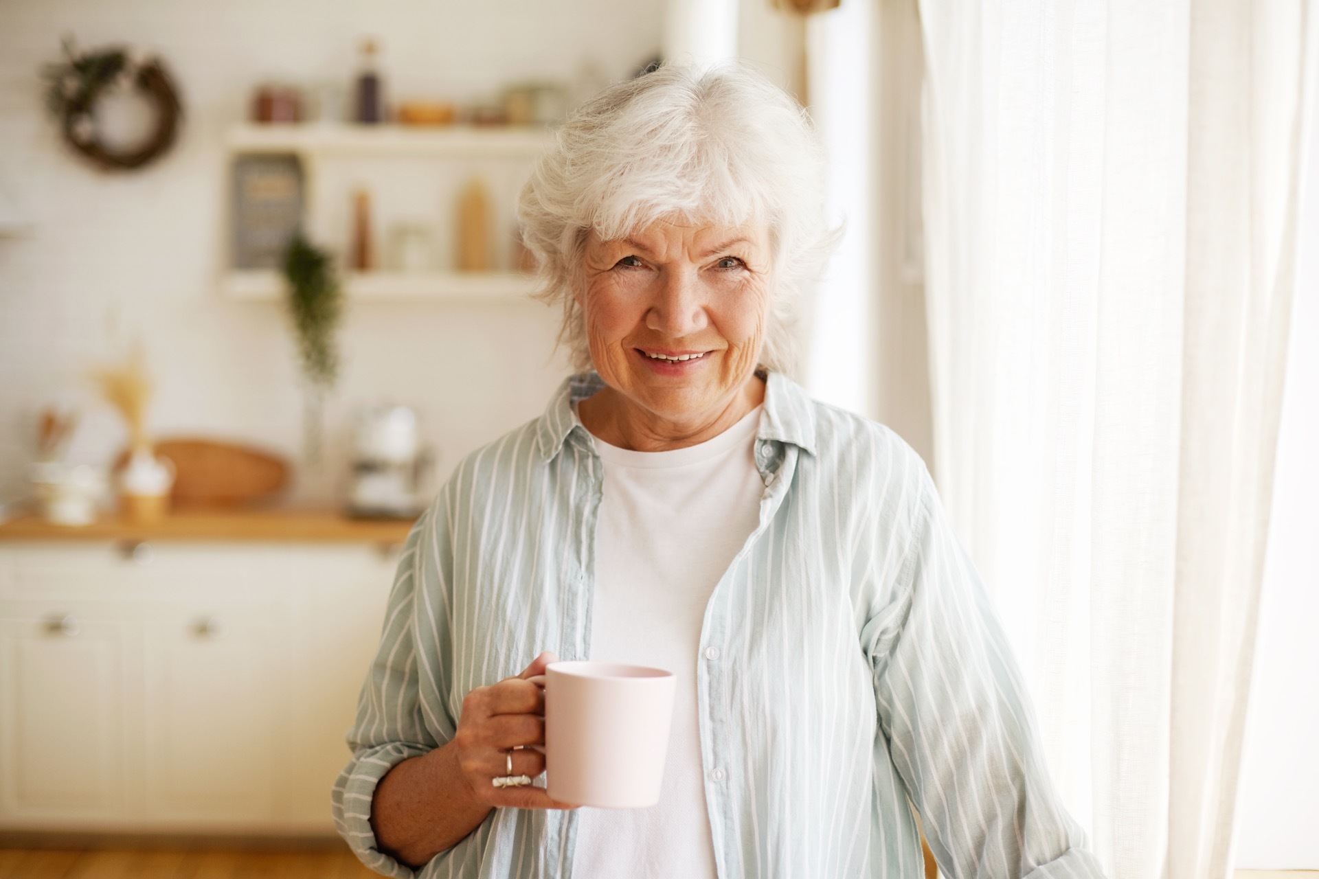 An older person, standing in a kitchen, holding a mug of drink and smiling at the camera; our Solicitors for the Elderly accredited Solicitor, Naomi Pinder, discusses care fees and future planning.